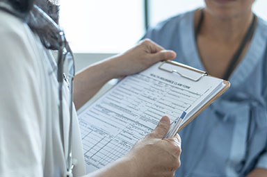 A doctor, with a stethoscope visible, holds a clipboard with medical forms while speaking to a patient.